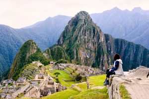 Mujer sentada junto a la casa del guardián en Machu Picchu, con la ciudadela y la montaña Huayna Picchu al fondo